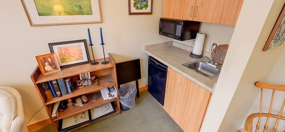 A small kitchenette area with a countertop, sink, microwave, and mini refrigerator. Next to it is a wooden bookshelf with framed photos, books, and decorative items. A wooden chair is partially visible on the right side.