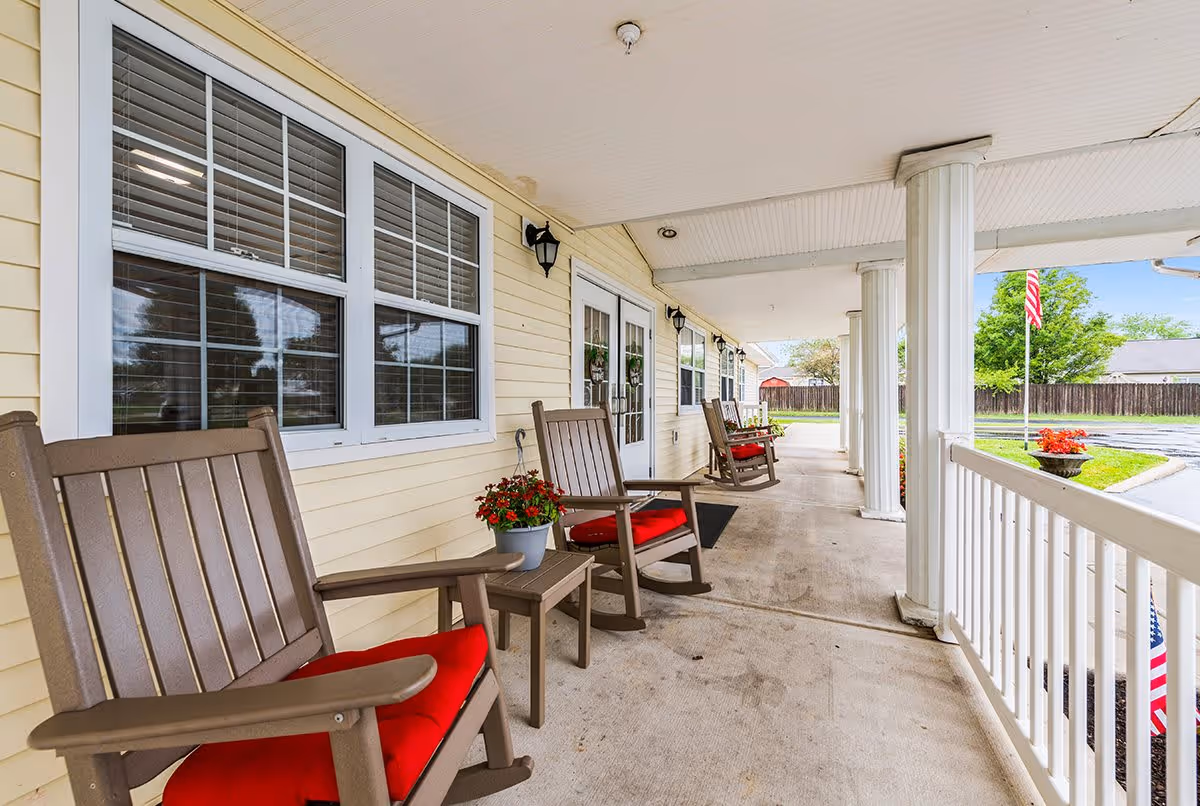 A covered porch area with beige siding and white trim, featuring several brown rocking chairs with red cushions and small tables with potted red flowers. White columns support the roof, and an American flag is visible in the background near a fenced yard.