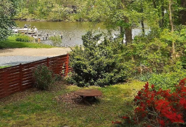 A peaceful outdoor scene featuring a grassy yard with a round wooden table, surrounded by green bushes and trees. In the background, there is a body of water with a dock and boats, and a red wooden fence partially enclosing the yard.