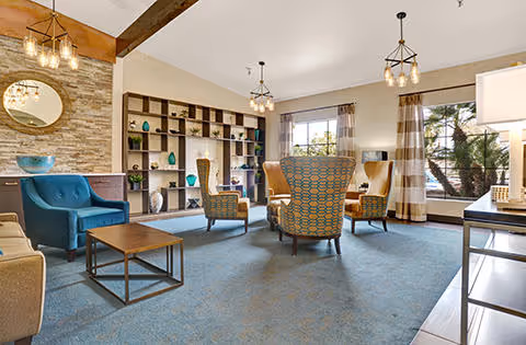 A spacious living room area with blue carpet, several armchairs including a blue one and patterned ones, a wooden coffee table, built-in shelves with decorative items, large windows with striped curtains, and modern hanging light fixtures.