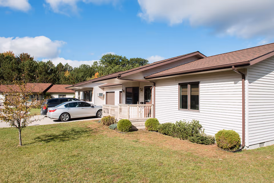 Single-story light-colored residential building with a small porch, shrubs, and two cars parked on a driveway under a blue sky.