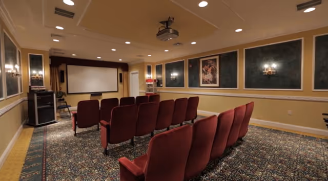 A small theater room with two rows of red cushioned chairs facing a large white projection screen. The room has patterned carpet, framed artwork on the walls, and a ceiling-mounted projector.