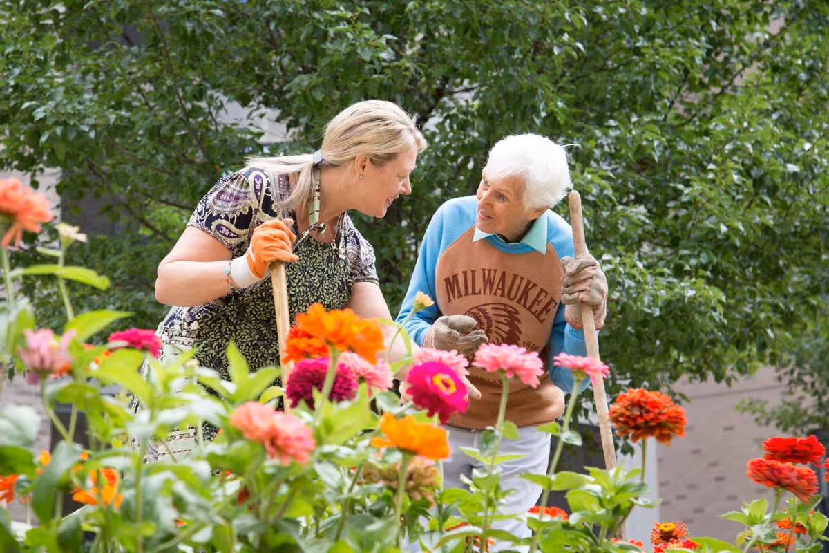 Two women gardening together outdoors surrounded by colorful flowers and green foliage. One woman is elderly with white hair wearing a Milwaukee sweatshirt and gloves, holding a gardening tool. The other woman has blonde hair tied back, wearing gardening gloves and an apron, leaning in to talk with the elderly woman.