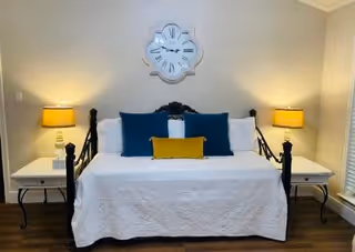 A bedroom with a black metal daybed covered with a white quilt and decorated with three pillows: two large blue ones and a smaller yellow one. On each side of the bed are white nightstands with yellow lamps. A large decorative wall clock hangs above the bed on a light-colored wall.