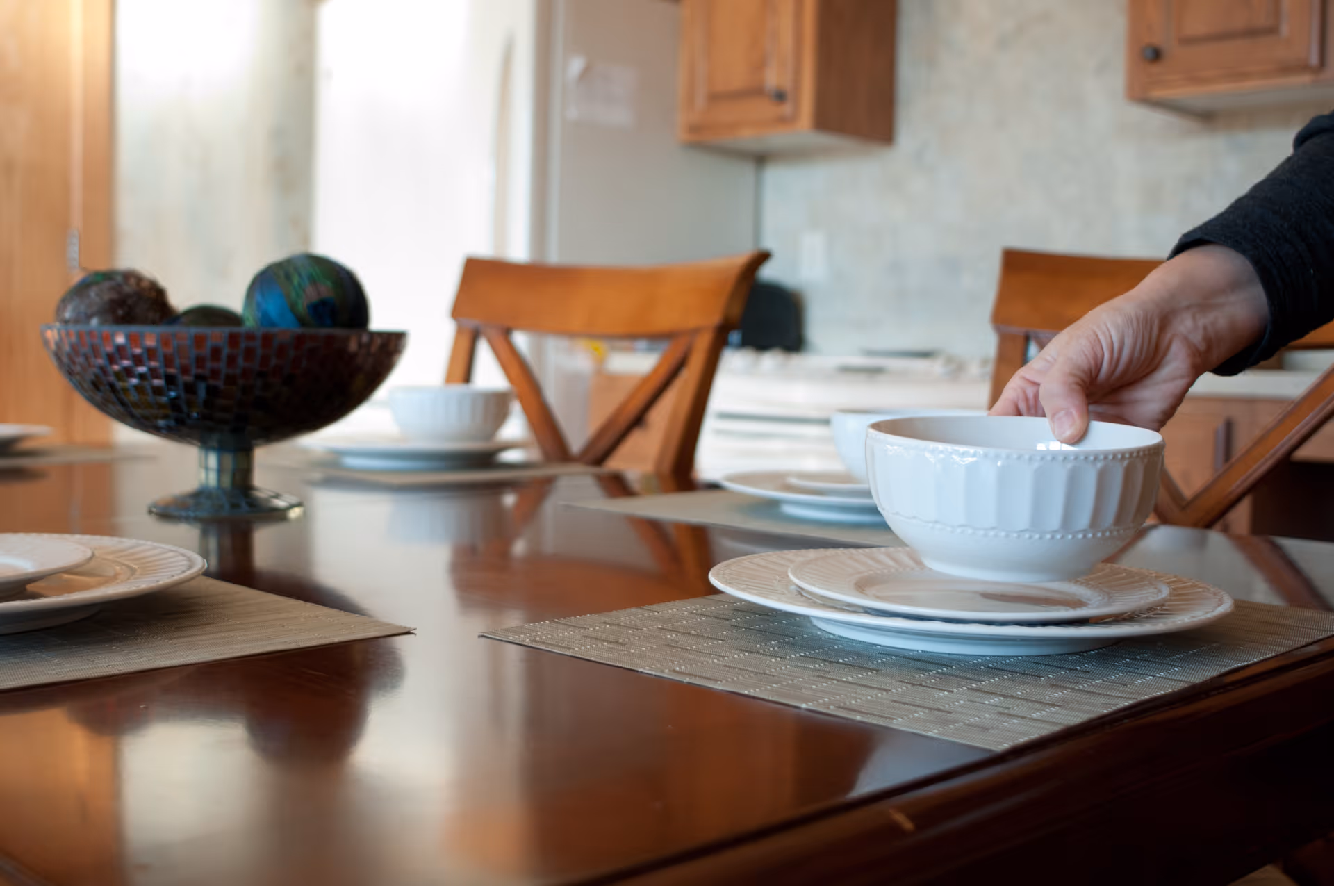 A close-up view of a dining table set with white plates and bowls on placemats. A hand is placing a white bowl on a plate. In the background, wooden chairs and kitchen cabinets are visible along with a decorative bowl filled with colorful spheres on the table.
