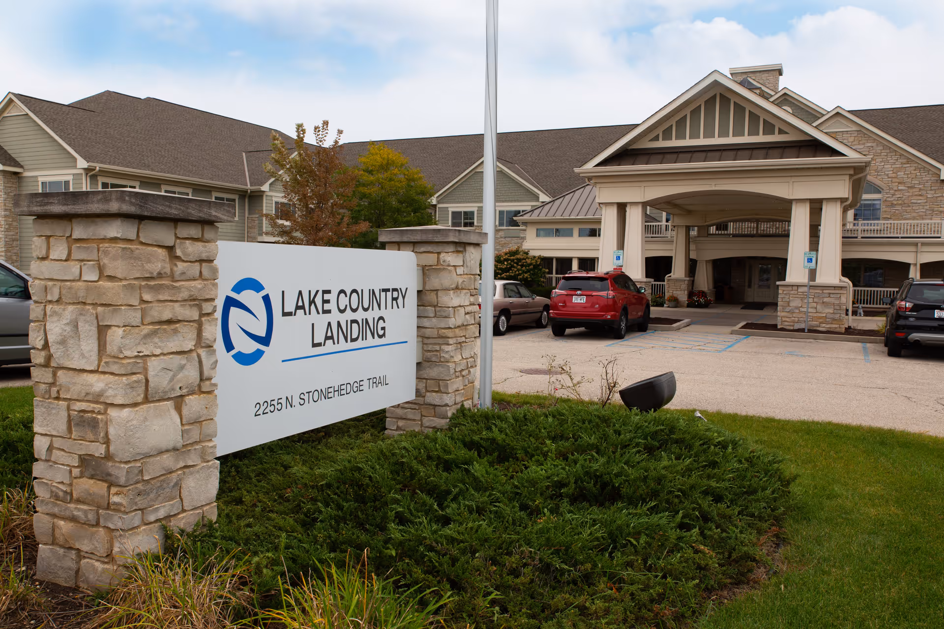 Exterior view of Lake Country Landing facility showing the main entrance with a covered drop-off area, parked cars, and a stone sign with the facility name and address 2255 N. Stonehedge Trail.