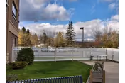 Outdoor area of a senior living facility with a green lawn, bushes, a white fence, a bench, and a paved walkway. Trees and a cloudy sky are visible in the background.