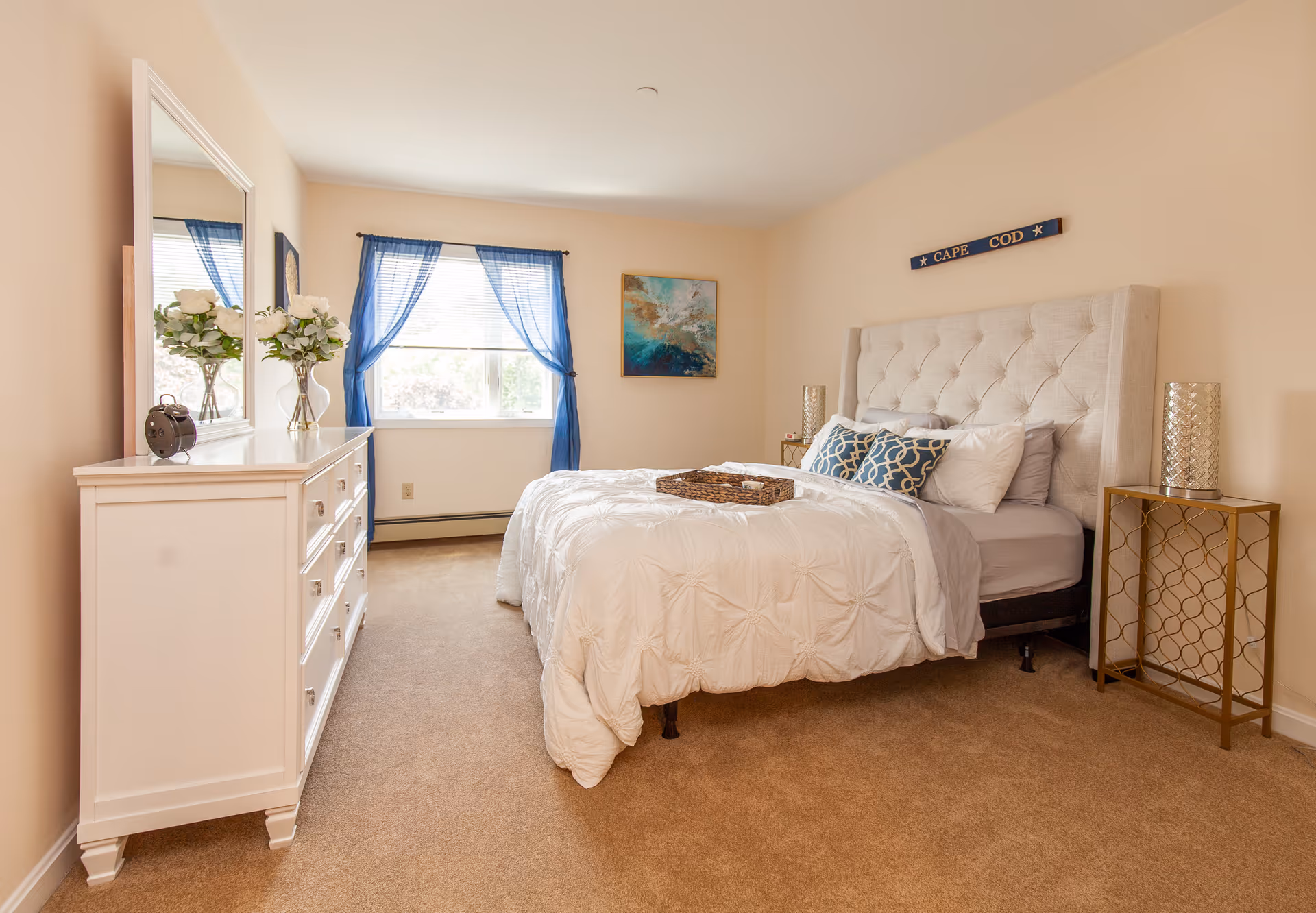 A cozy bedroom with a beige tufted headboard bed dressed in white bedding and decorative pillows. There is a white dresser with a mirror and a vase of flowers on top to the left, and a window with blue curtains letting in natural light. A small gold side table with a decorative lamp is on the right side of the bed. The walls are painted light beige, and a piece of artwork hangs above the bed.