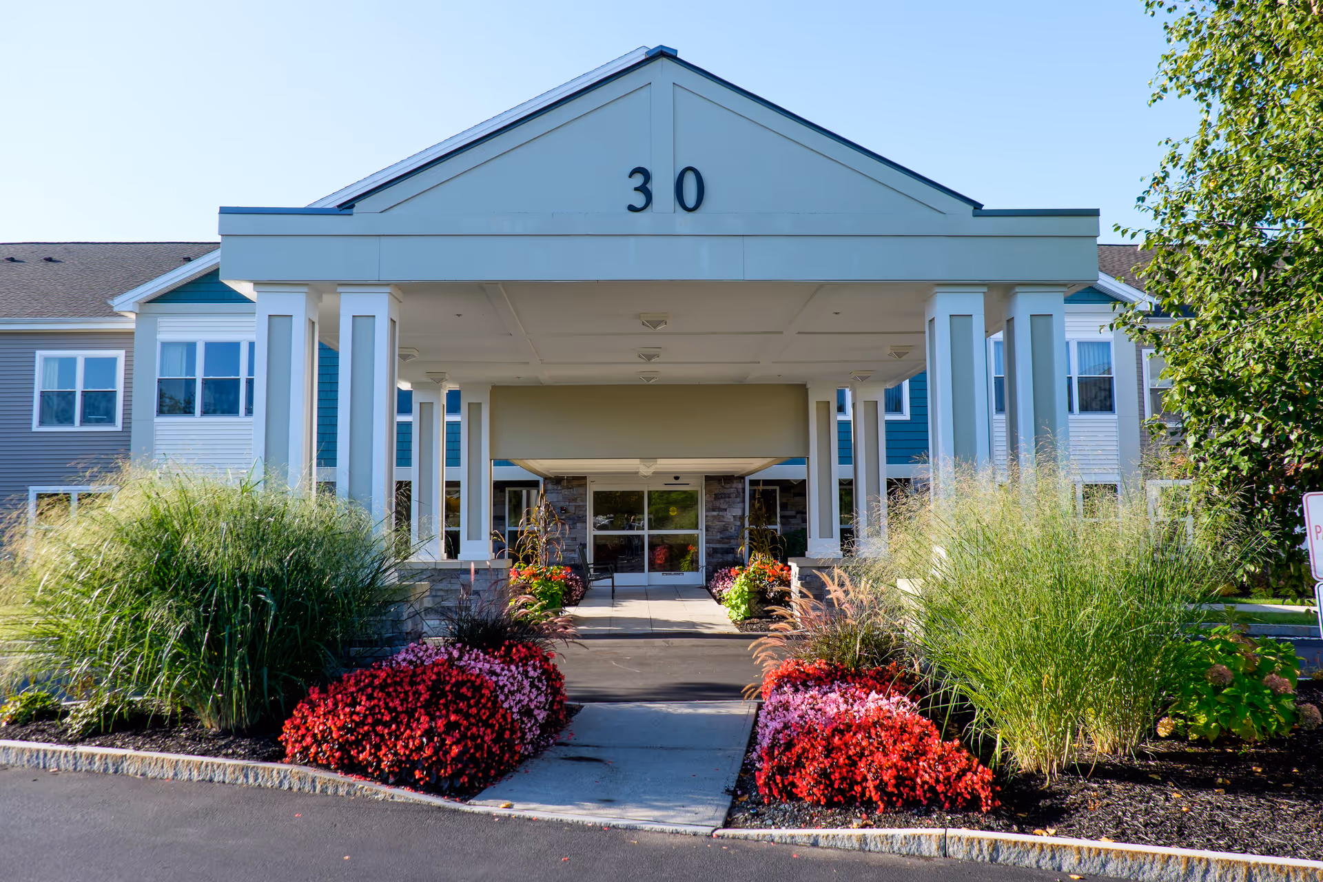 Entrance of a senior living facility with a covered driveway supported by white columns, landscaped with green bushes and red flowers on either side. The building has a blue and white exterior with multiple windows and the number 30 displayed prominently above the entrance.