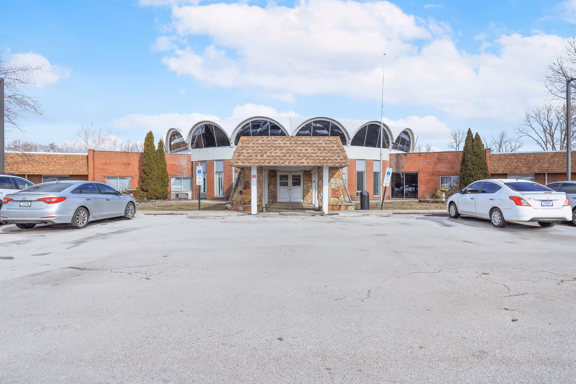 Front exterior view of Envive Healthcare of Indianapolis building with a unique curved roof design, a covered entrance, two parked cars, and a clear sky with some clouds.