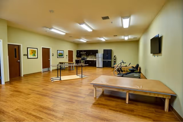 A spacious room with wooden flooring and pale yellow walls featuring exercise equipment including a stationary bike and treadmill, a large padded therapy table, parallel bars for walking exercises, and a kitchenette area with cabinets, a refrigerator, and a microwave. There are three closed doors and framed artwork on the walls, with a flat-screen TV mounted on the right wall.