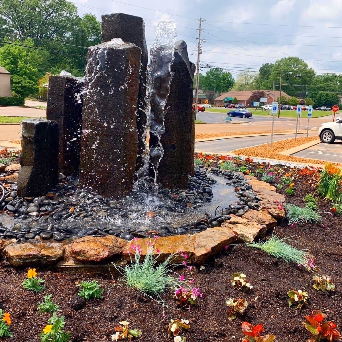 Outdoor water fountain with multiple vertical stone pillars with water flowing down, surrounded by a circular arrangement of rocks and a flower bed with various plants and flowers. In the background, there is a parking lot with cars and trees under a partly cloudy sky.