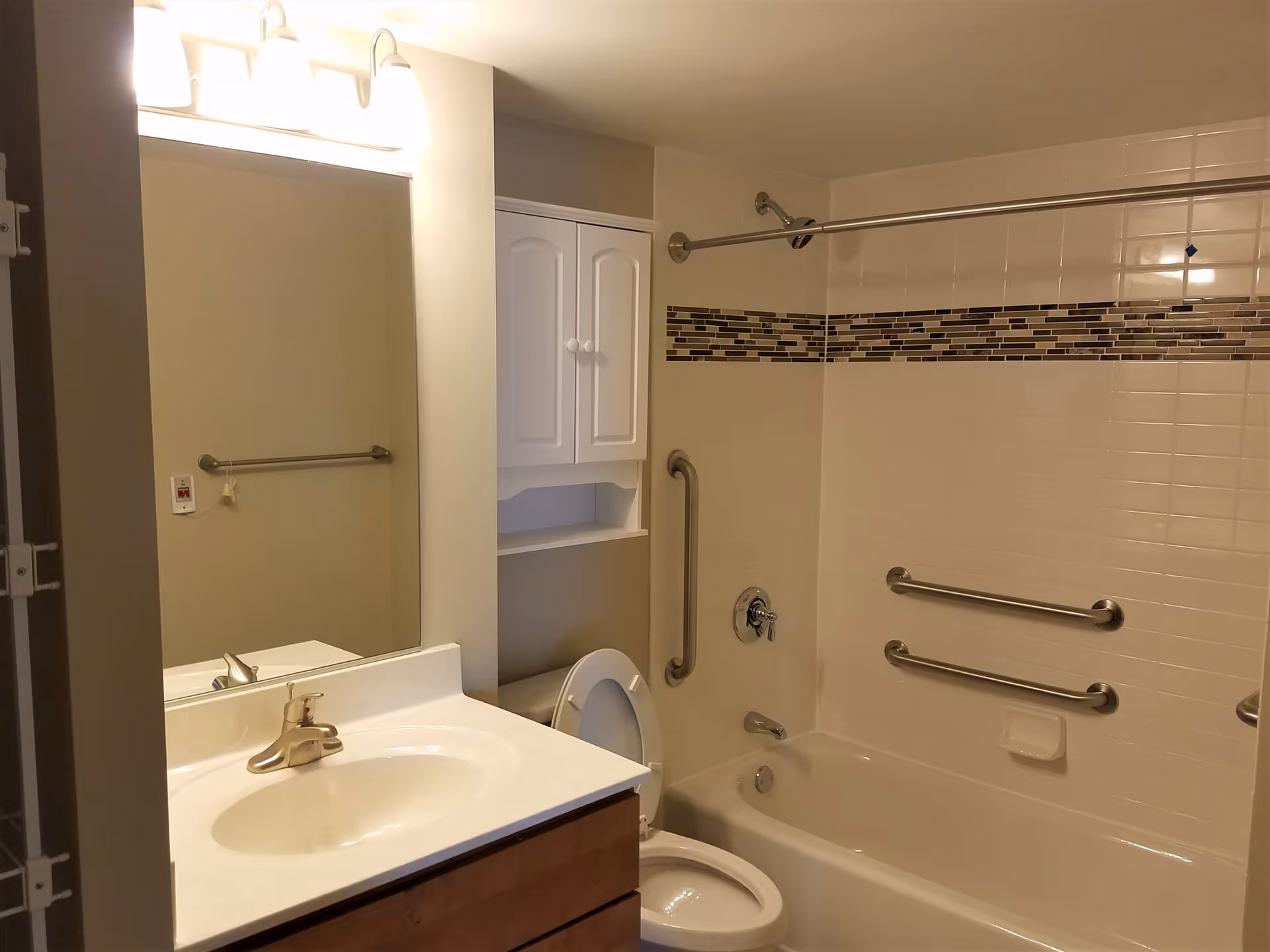 A bathroom with a white sink and countertop, a large mirror above the sink, a toilet with the lid open, and a bathtub with a shower. The bathtub area has white tiles with a decorative horizontal strip of smaller dark and light tiles. There are three metal grab bars installed around the bathtub for safety. A white cabinet is mounted on the wall above the toilet. The lighting fixture above the mirror has three lights.
