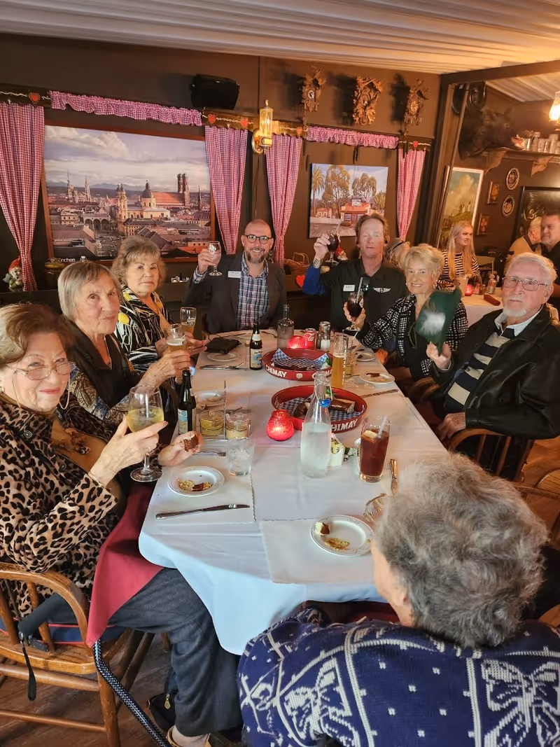 A group of elderly people and two staff members sitting around a dining table in a cozy restaurant setting, raising their glasses in a toast. The table is set with drinks, plates, and candles. The room is decorated with paintings, red checkered curtains, and wooden wall ornaments.