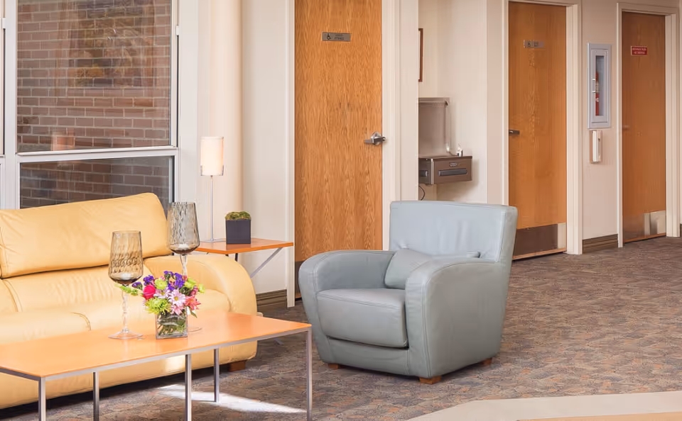 A cozy seating area in a hallway with a beige leather sofa, a light blue armchair, a wooden coffee table with two decorative glasses and a small flower arrangement, a side table with a small plant and lamp, and wooden doors in the background including an accessible restroom door and a water fountain.