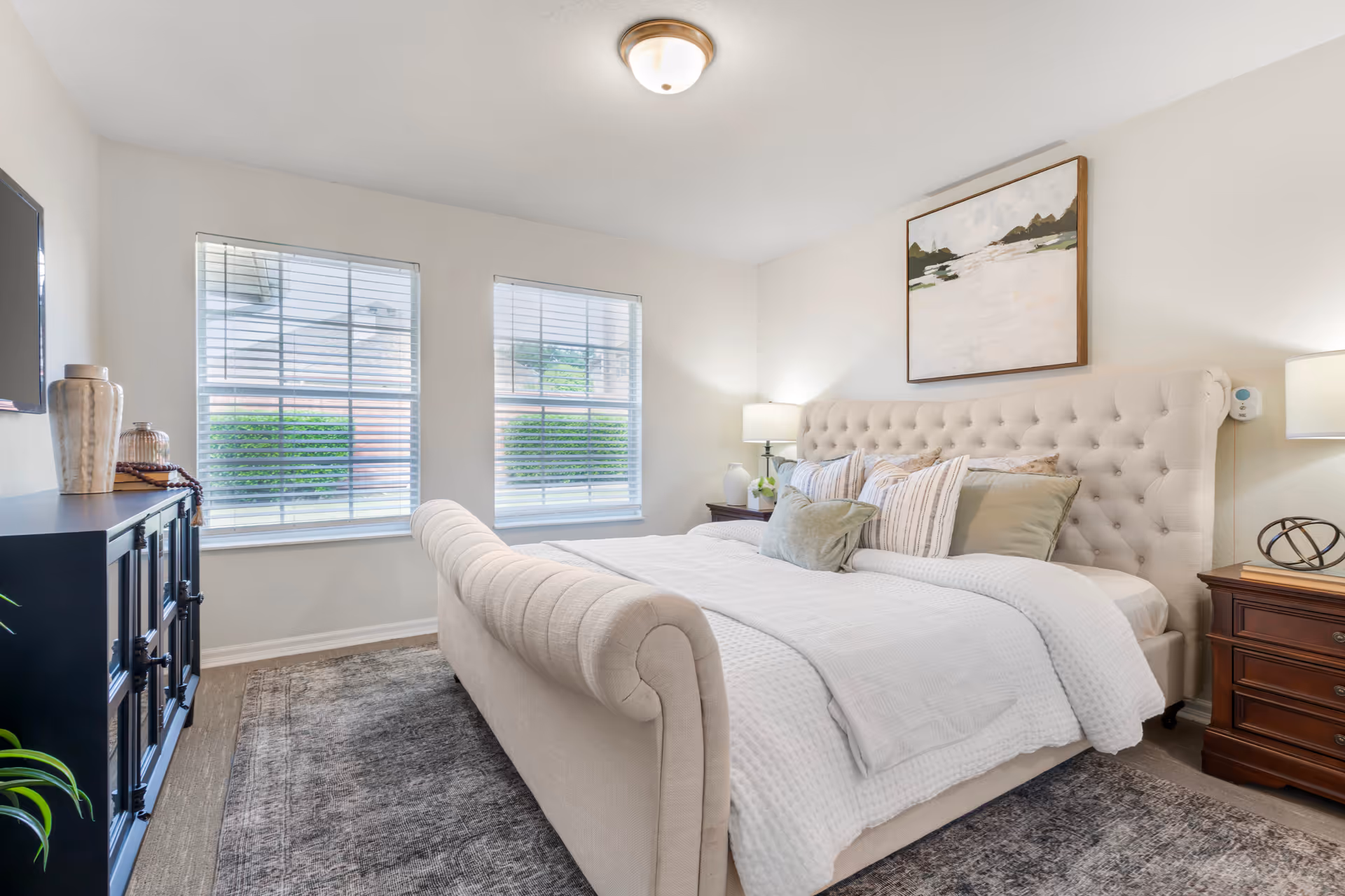 A bright and cozy bedroom with a large tufted beige bed adorned with multiple pillows and a white textured blanket. Two windows with white blinds allow natural light to fill the room. On the right side, there is a wooden nightstand with a lamp and decorative items. A dark cabinet with vases and decor is on the left side, and a framed abstract painting hangs above the bed.