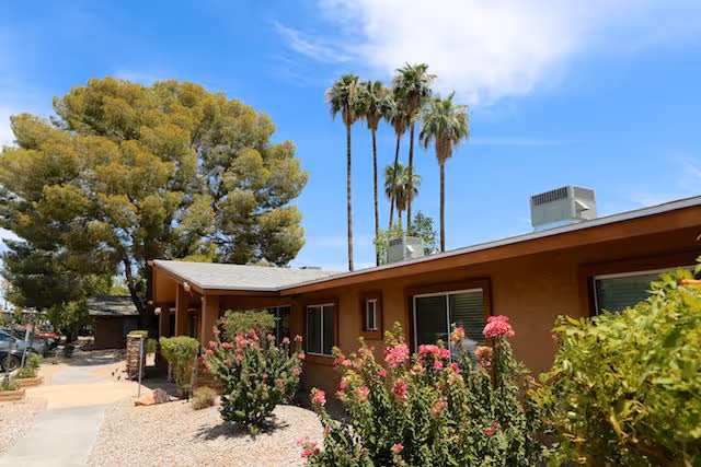Exterior view of a single-story building with brown walls and several windows, surrounded by desert landscaping with flowering bushes, palm trees, and other greenery under a blue sky with some clouds.