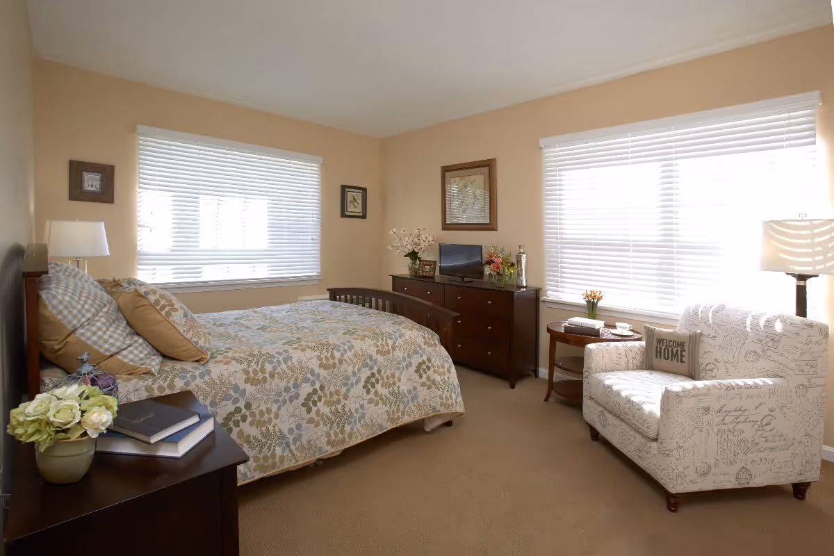 A cozy bedroom with a bed covered in a floral patterned quilt and multiple pillows. There is a wooden nightstand with a lamp, books, and a flower pot. Across from the bed is a dark wooden dresser with a small TV, flowers, and decorative items. Next to the dresser is a round side table with a small flower vase and a white armchair with script patterns and a 'Welcome Home' pillow. The room has beige walls and two large windows with white blinds letting in natural light.