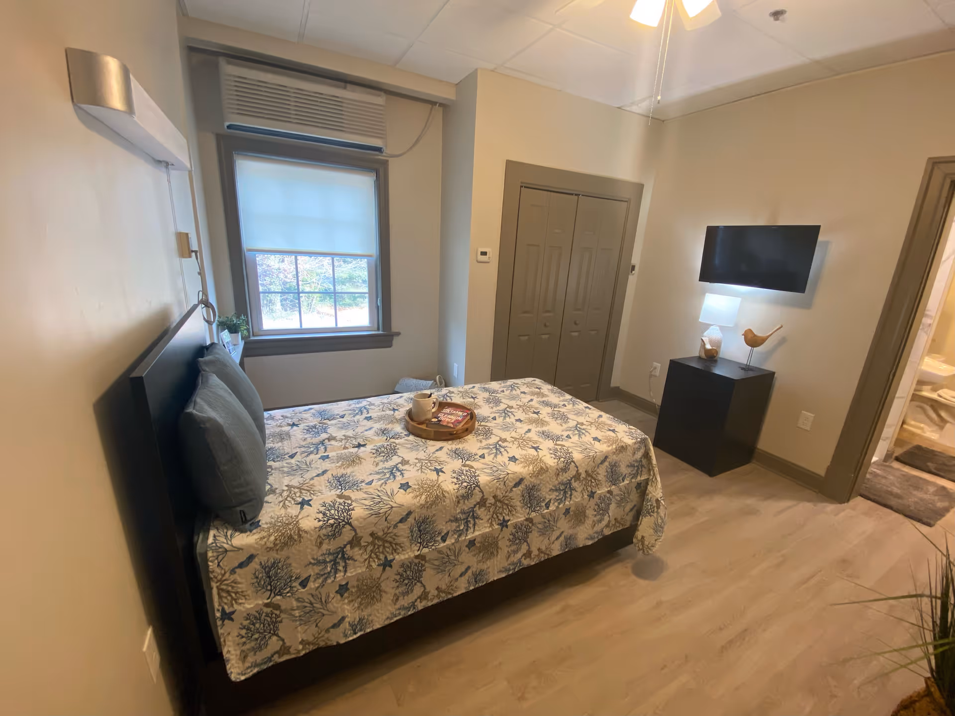 Small furnished bedroom with a single bed, window, wall-mounted TV, and nightstand.