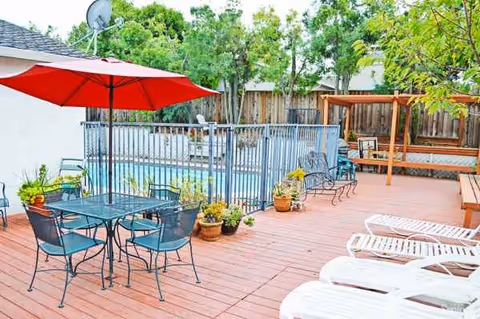 Outdoor patio at a senior care facility with tables, chairs, umbrellas, lounge chairs, and a pool.