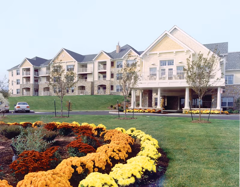 Front exterior of a multi-story senior living building with a covered entrance, balconies, and colorful flower beds in the foreground.