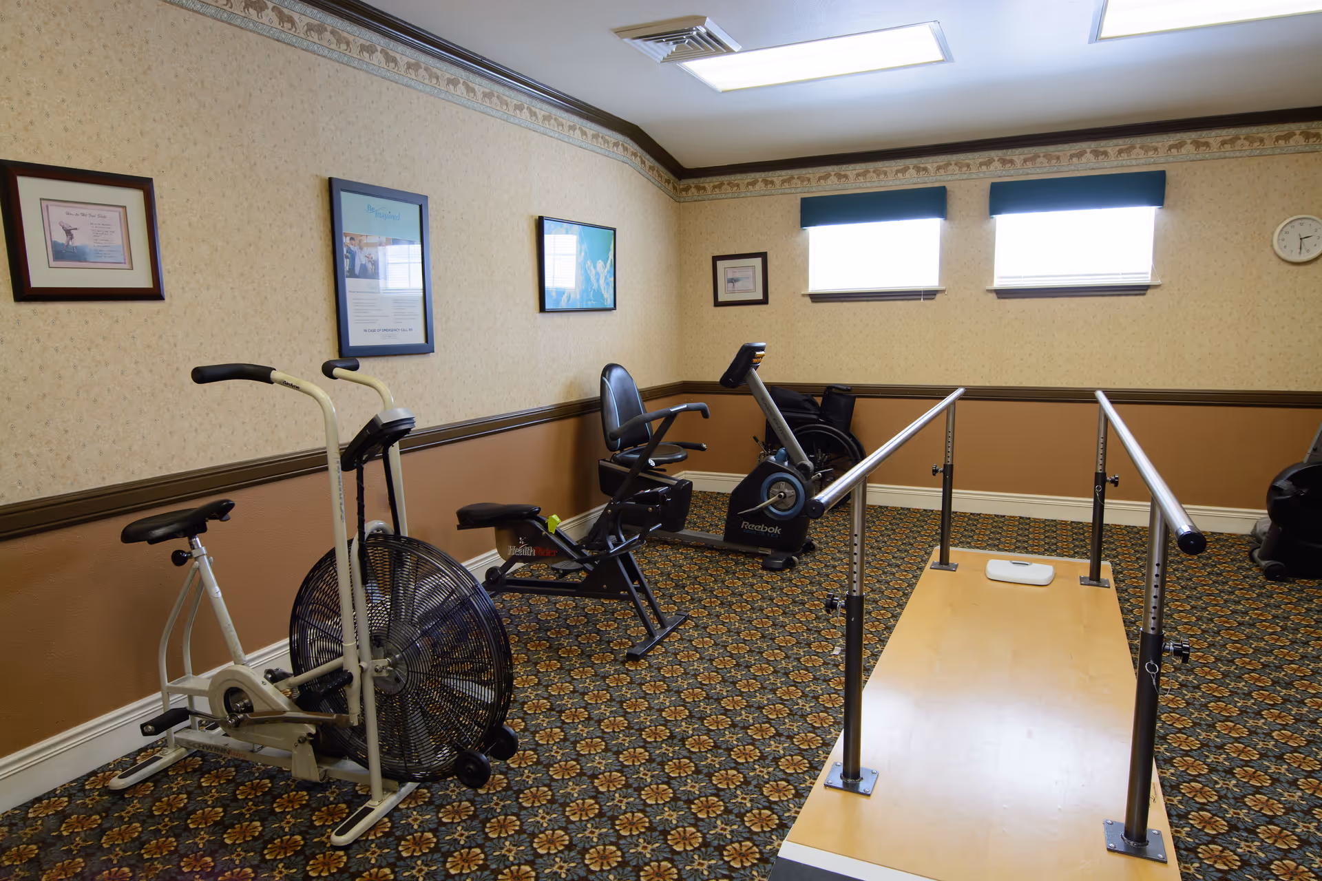 A small exercise room with patterned carpet and beige walls featuring three framed pictures. The room contains exercise equipment including a Schwinn stationary bike, a HealthRider recumbent bike, a Reebok exercise bike, and parallel bars for walking support. Two windows with blinds and a clock are on the far wall.