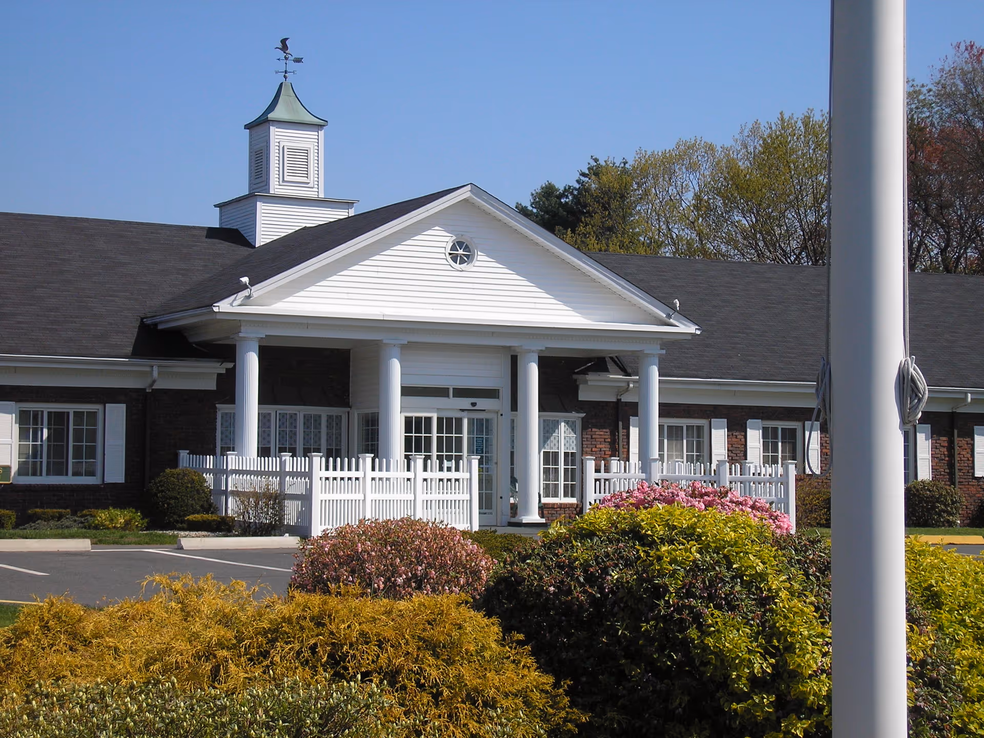 Front exterior view of a single-story building with white columns, a small cupola with a weather vane on the roof, white fencing, and landscaped bushes and flowers in front under a clear blue sky.