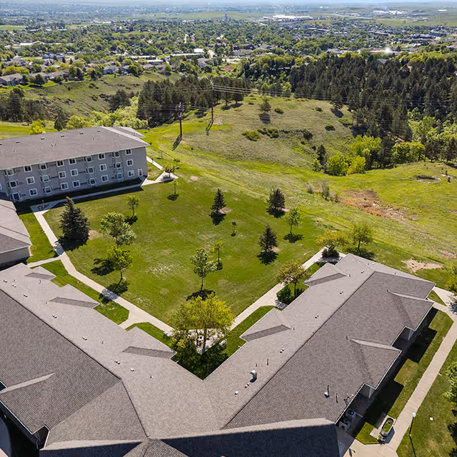 Aerial view of Good Samaritan Society - Echo Ridge - The Manor showing multiple connected buildings surrounding a large green courtyard with scattered trees and walking paths. Beyond the facility, there is a mix of open grassy areas, trees, and a distant view of a residential neighborhood and fields.