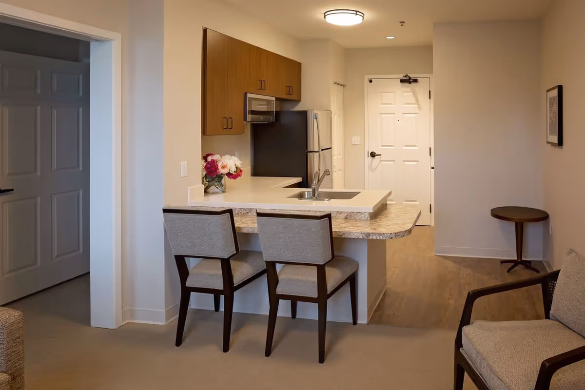Interior view of a senior living facility kitchen and living area at Jubilee House. The kitchen features a countertop with two chairs, a sink, a refrigerator, a microwave, and wooden cabinets. Adjacent to the kitchen is a small living area with a cushioned chair and a small round side table against the wall. The entrance door is visible in the background.