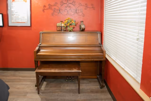 A wooden upright piano with a matching bench in front of a red wall. On top of the piano are decorative items including a vase with flowers and two other ornamental containers. To the right is a window with white blinds partially closed. The floor is wooden.