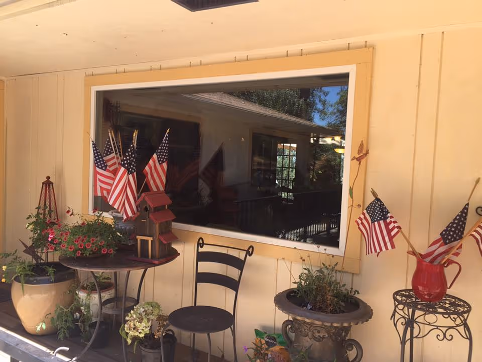 A house porch with potted plants, a chair, a small table and decorative American flags in front of a large window.