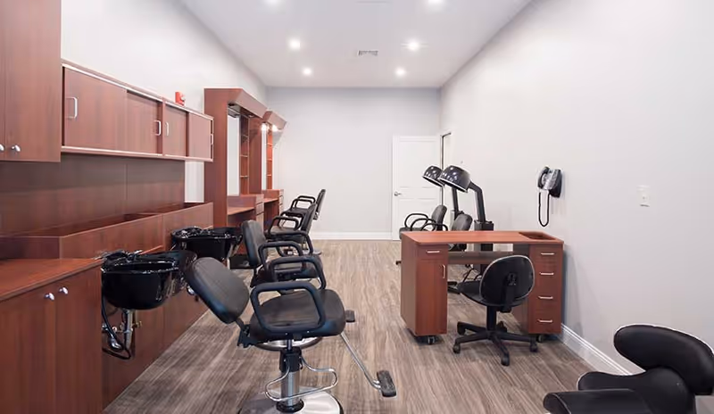 Interior view of a salon area with multiple black salon chairs, hair washing sinks, wooden cabinetry, and hair drying stations. The room has light gray walls, wood flooring, and recessed ceiling lights.
