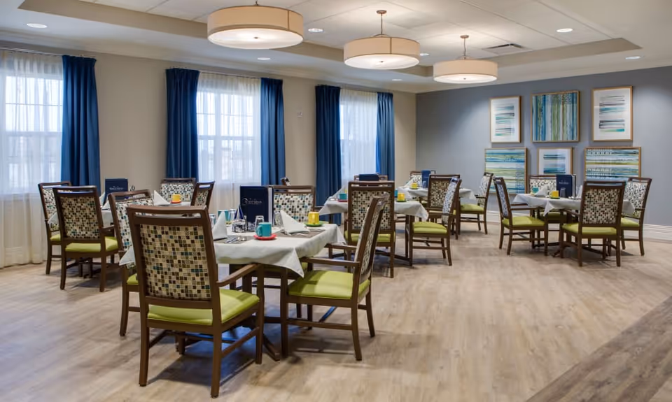 A bright dining room with several tables covered in white tablecloths, each set with colorful cups, napkins, and menus. The room has wooden chairs with patterned backs and green cushions, large windows with sheer white curtains and dark blue drapes, light wood flooring, and modern circular ceiling lights. The walls are decorated with a series of abstract blue and green paintings.