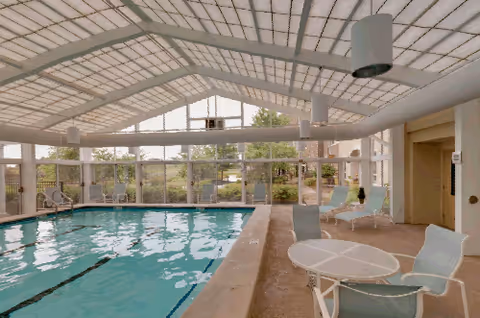 Indoor swimming pool area with large windows and a glass ceiling allowing natural light. The pool has lane markings and is surrounded by a tiled deck with white patio tables and chairs. There are some lounge chairs near the pool and greenery visible outside through the windows.