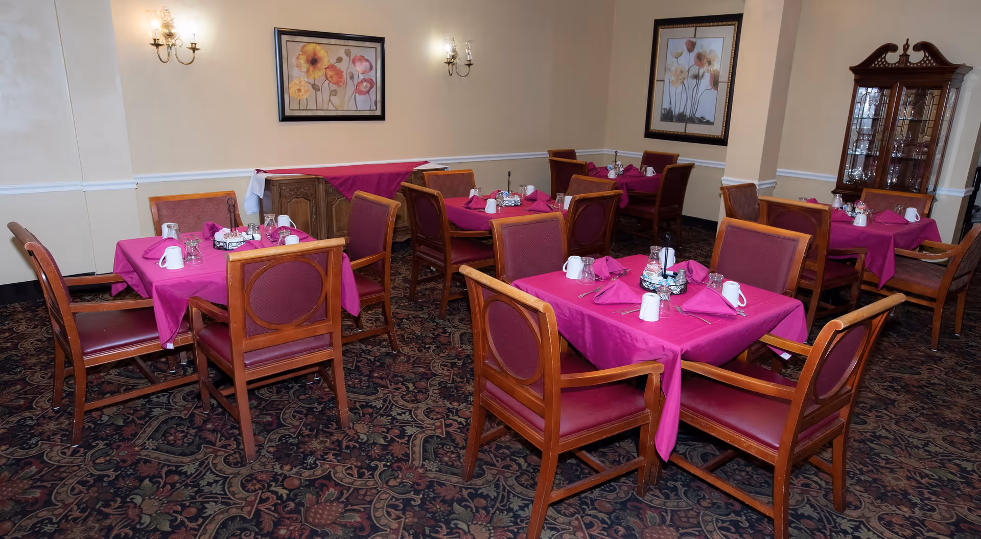 Dining room with several wooden tables covered with magenta tablecloths, each set with white mugs, glasses, and folded napkins. The room has patterned carpet, beige walls with floral paintings, wall sconces, and a wooden cabinet with glassware.
