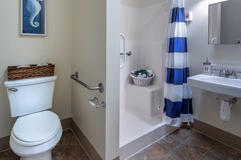 A clean bathroom featuring a white toilet with a wicker basket on top, a wall-mounted grab bar, a shower area with a blue and white striped curtain, a white sink with a soap dispenser, and a mirror above the sink. The floor is tiled in brown tones and there is a framed seahorse picture on the wall.
