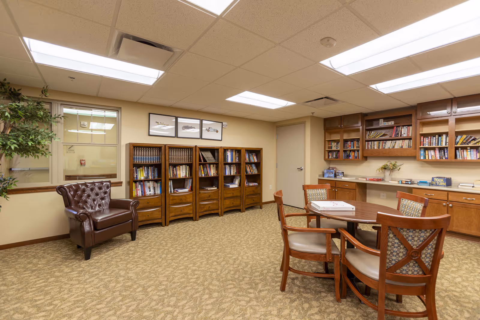 A cozy senior living common room with bookshelves, a round table and chairs, and a leather armchair.