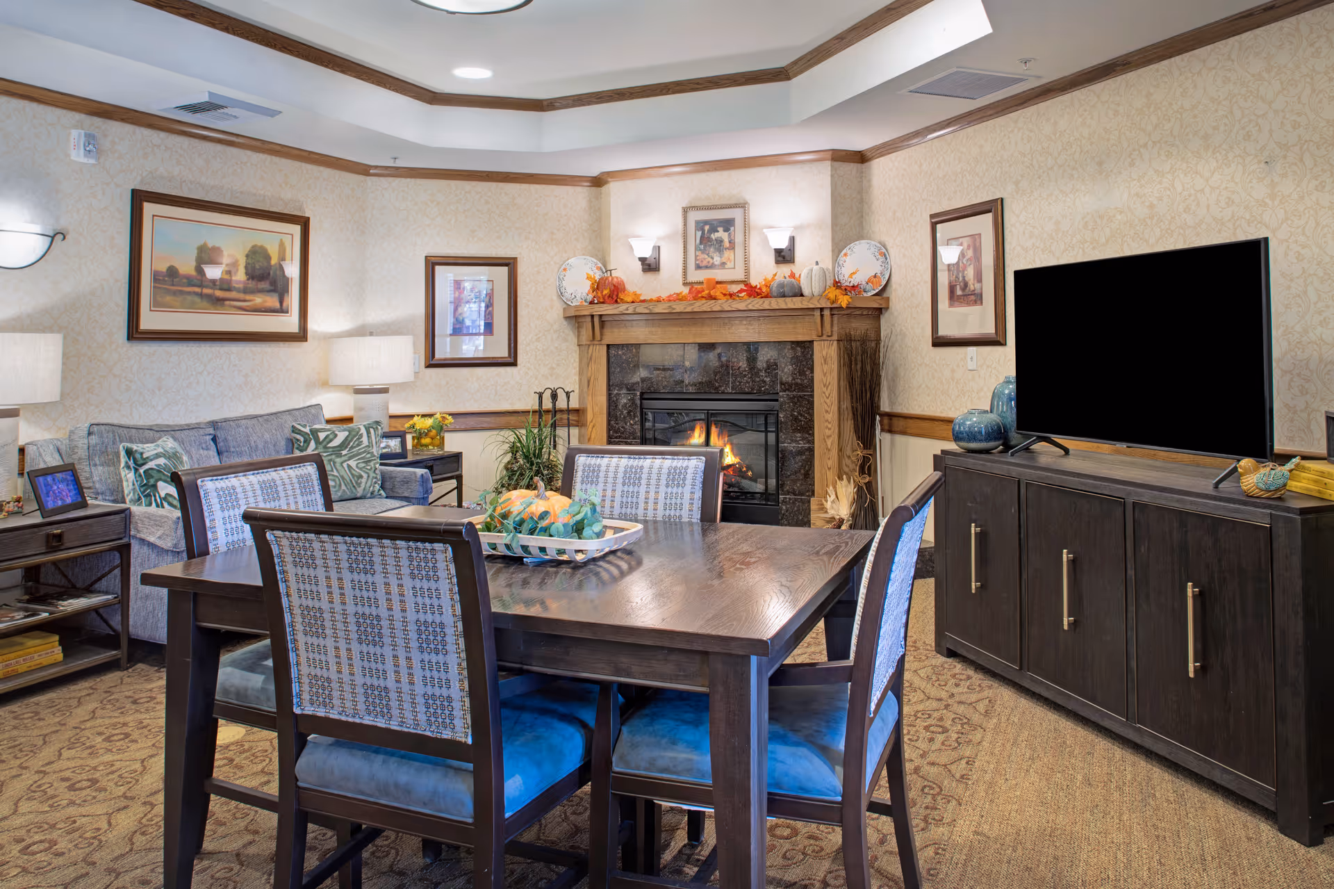 A cozy living room area with a wooden dining table and four chairs in the foreground. Behind the table is a fireplace with a wooden mantle decorated with autumn-themed items including pumpkins and leaves. To the left is a blue sofa with patterned cushions and side tables with lamps and framed pictures. On the right side, there is a dark wooden cabinet with a large flat-screen TV on top. The walls have a light patterned wallpaper and framed artwork.
