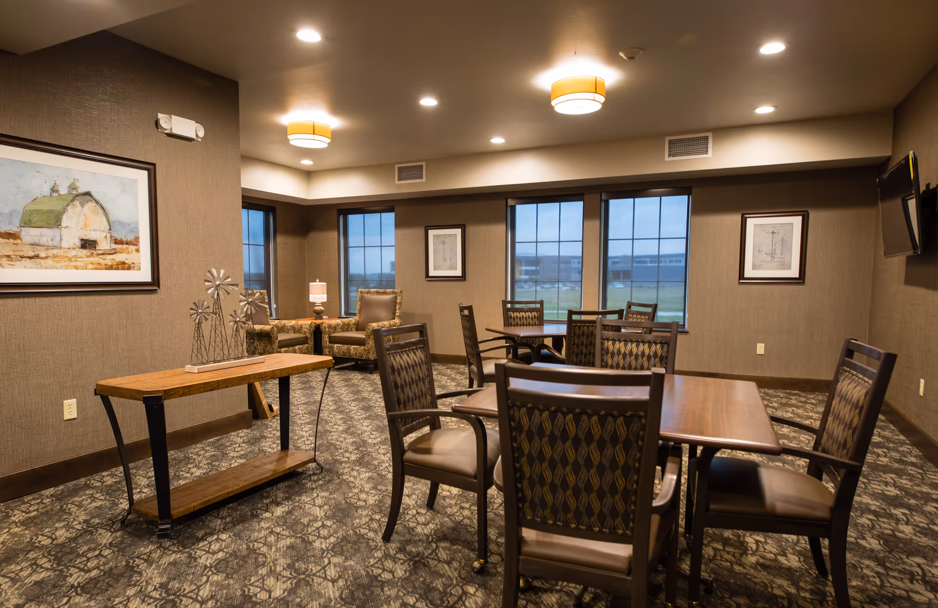 Well-lit senior living common room with several tables and chairs, armchairs near windows, and framed artwork on the walls.