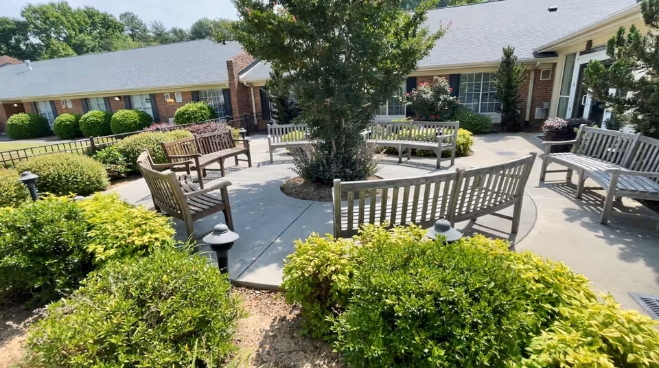 Sunny courtyard with a circular paved seating area and wooden benches surrounded by shrubs in front of a brick senior living building.