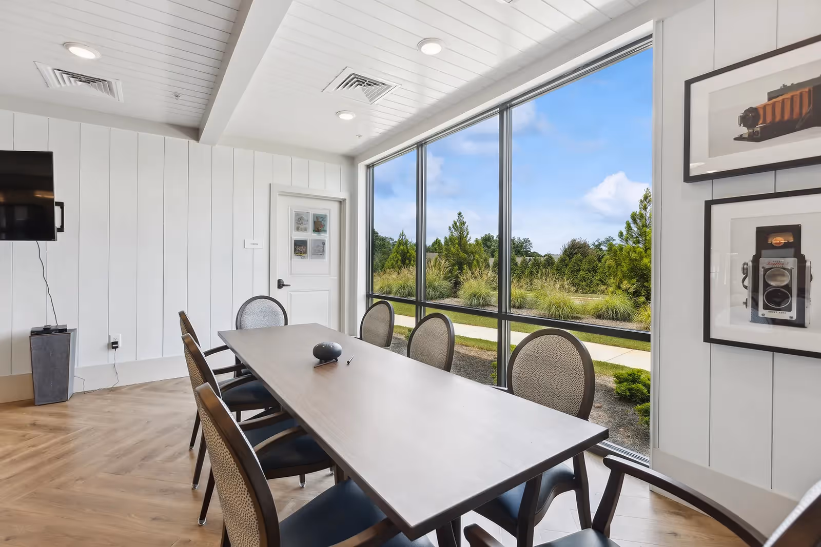 A bright meeting or dining room with a long rectangular table surrounded by six chairs. Large floor-to-ceiling windows offer a view of greenery and a walkway outside. The room has white paneled walls, a wall-mounted TV, and framed pictures of vintage cameras on one wall.