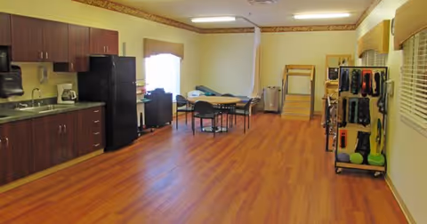 Large multipurpose activity room with a kitchenette on the left, a table and chairs in the center, and therapy/exercise equipment along the right wall.