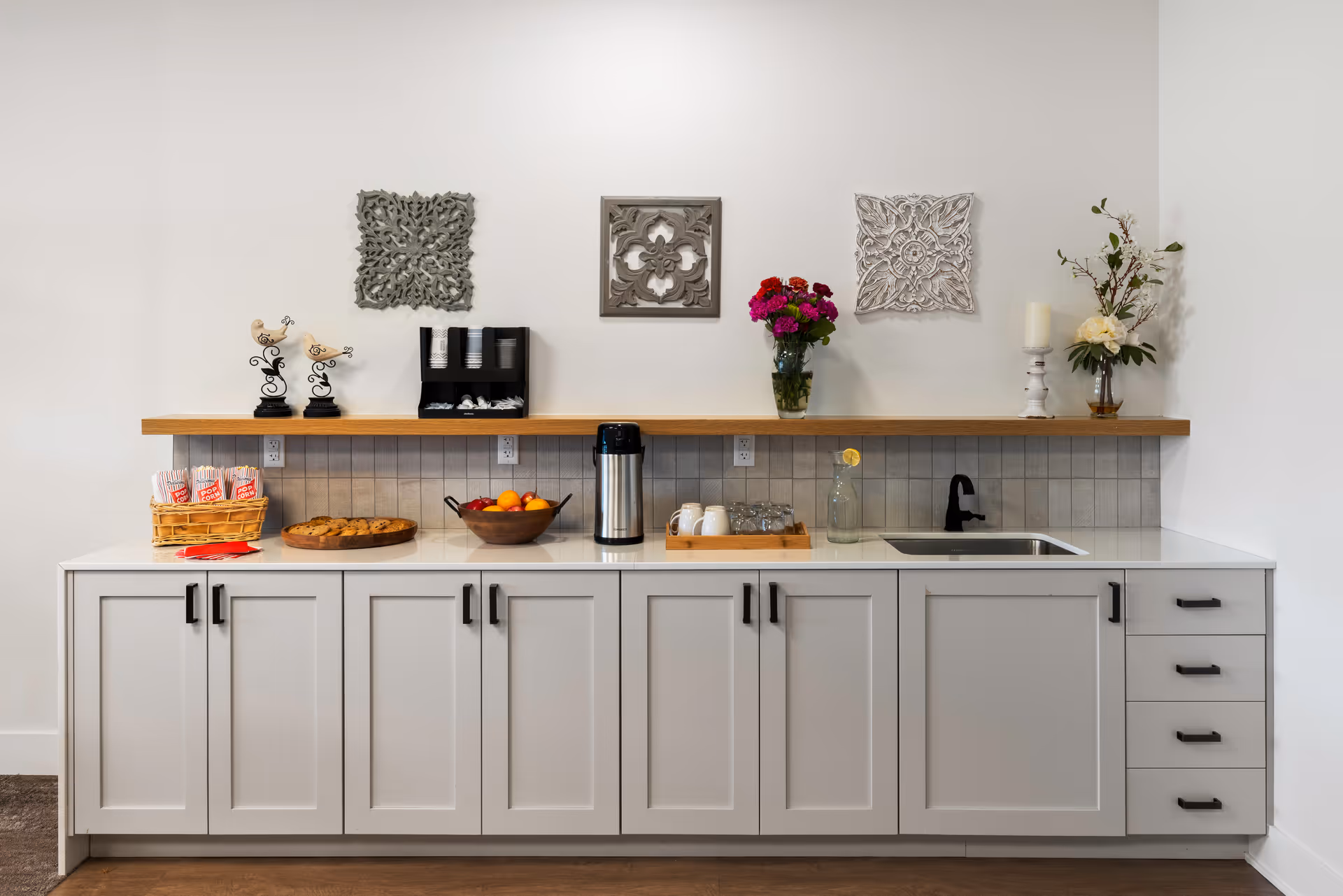 Well-lit kitchenette with white cabinets and a countertop holding a coffee urn, fruit bowl, pastries, sink, and decorative wall art.