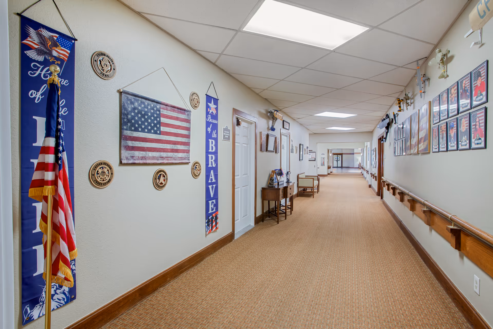 A long hallway in a senior living facility decorated with patriotic American flags, banners, and plaques on the walls. The hallway has beige carpet, handrails on both sides, and framed pictures and trophies displayed along the walls. There are chairs and small tables placed intermittently along the corridor.