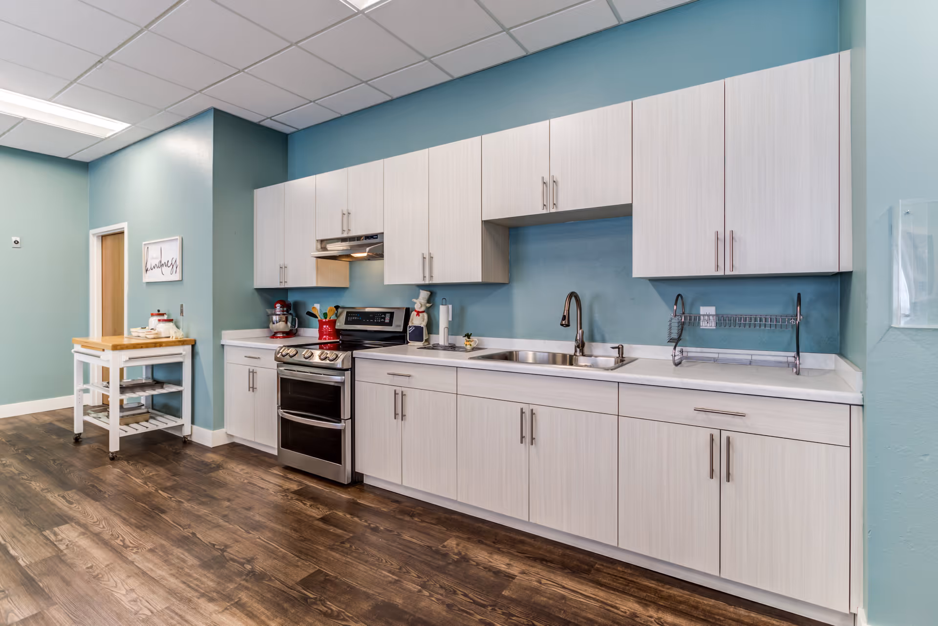 Communal kitchen with white cabinets, a stainless steel stove and double sink set against teal walls and wood-look flooring.
