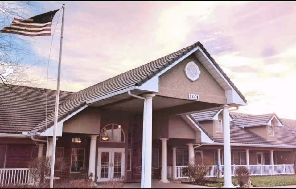 Exterior view of a building with a peaked roof and white columns at the entrance. An American flag is flying on a flagpole to the left. The building has multiple windows and a porch with white railings. The address number 5201 is displayed above the entrance.