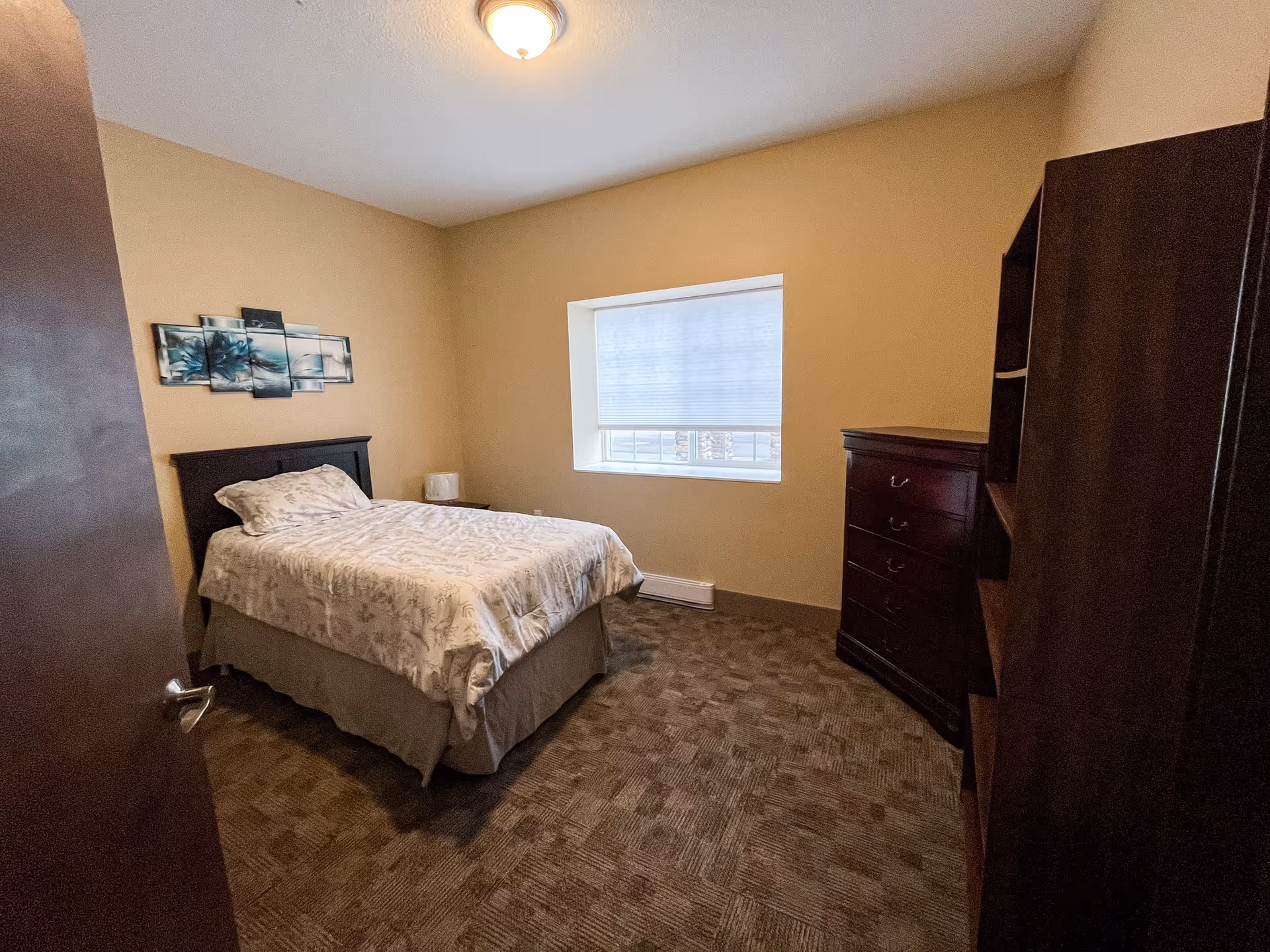 Simple furnished bedroom with a single bed, dresser, window with a blind, and artwork on a beige wall.