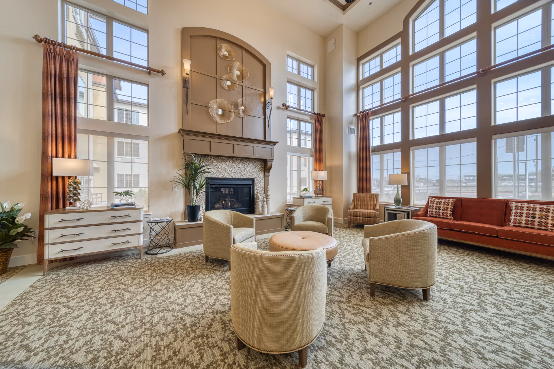 A spacious senior living facility common area with large floor-to-ceiling windows allowing natural light to fill the room. The room features a fireplace with decorative wall art above it, four beige armchairs arranged around a round ottoman, a red sofa with patterned pillows, two side tables with lamps, and tall curtains framing the windows. The carpet has a patterned design, and there are plants and decorative items on dressers near the windows.