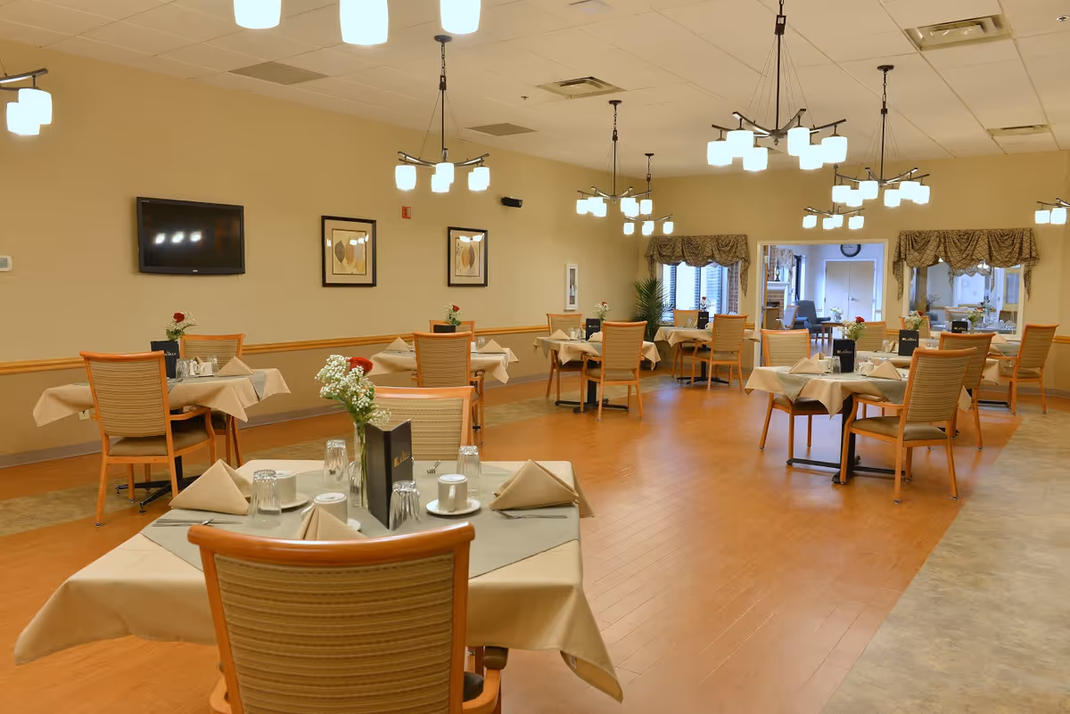Well-lit dining room with multiple set tables and chairs, hanging light fixtures, and a wall-mounted TV.