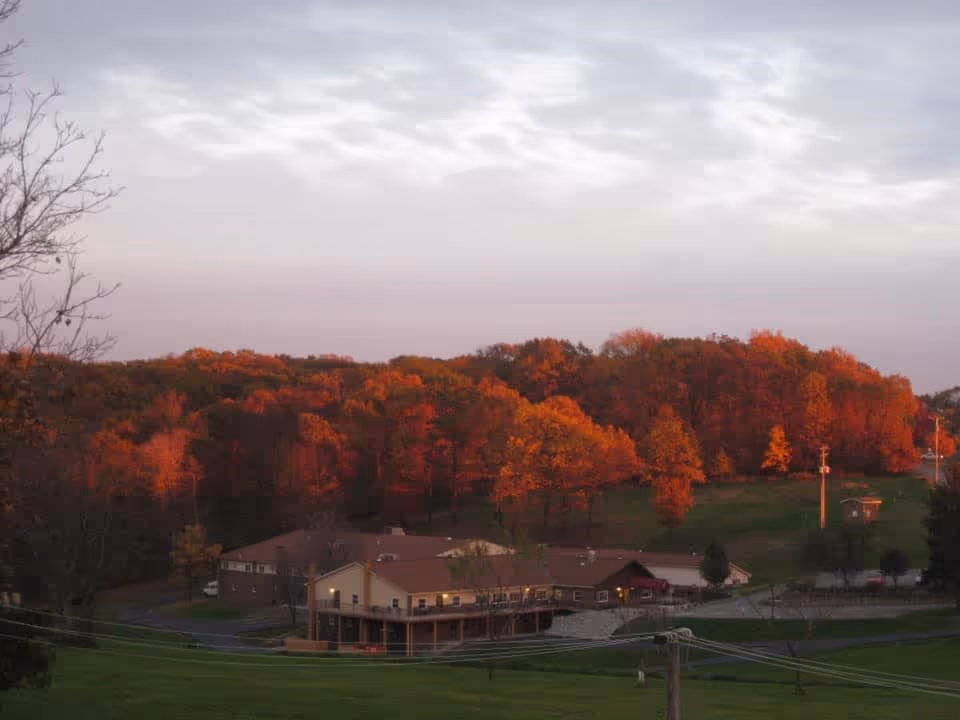 A scenic view of a senior living facility named Elmcroft surrounded by trees with autumn foliage in shades of orange and red under a cloudy sky during sunset or sunrise.